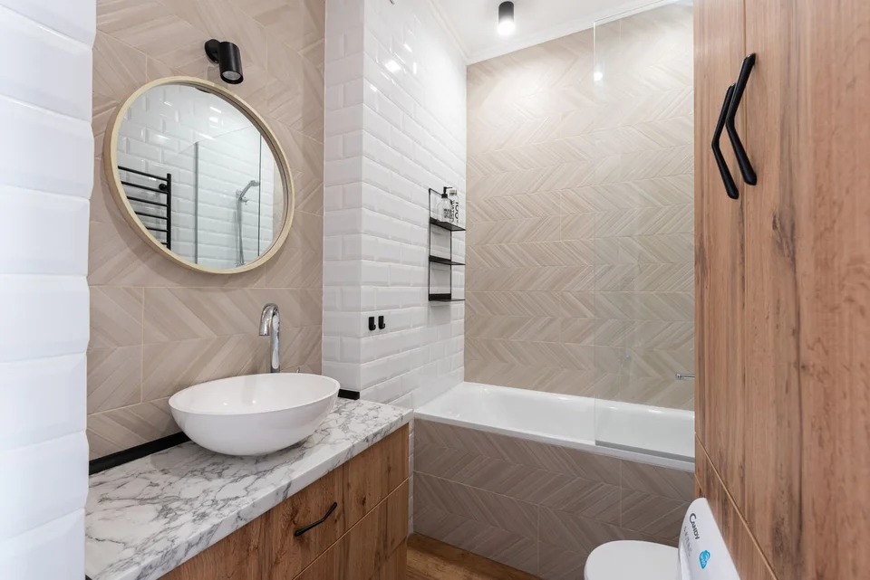 Stock bathroom reference with wood cabinets, a round mirror, white basin and beige herringbone bath tiles